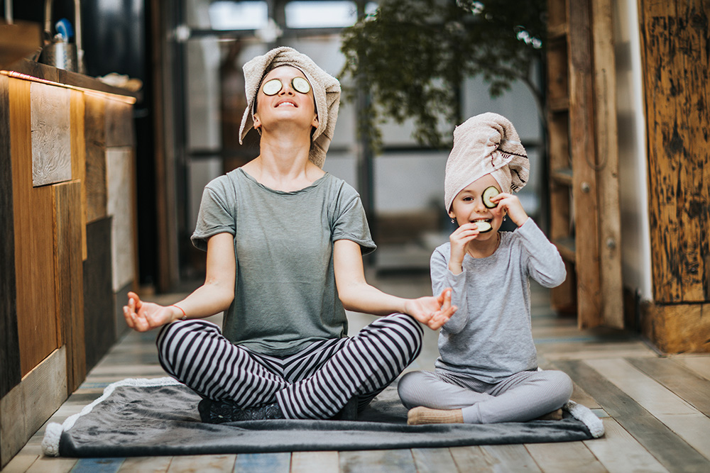 Mother and little girl taking care of their bodies in the morning while doing Yoga meditation exercises at home. Girl is cheating while eating cucumbers from her eyes and looking at camera.